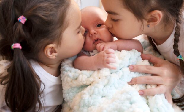 Harmonie ohne Geschwisterrivalität: Zwei ältere Schwestern küssen ein Baby.